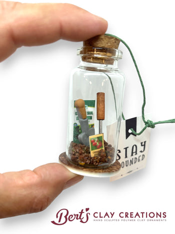 Glass bottle with cork lid containing miniature gardening tools and soil, held by a hand. 'Stay Grounded' label and 'Bert's Clay Creations' branding.
